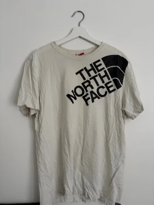 Vit t-shirt från The North Face - Säljer en vit t-shirt från The North Face med stor svart logga tryckt diagonalt över bröstet och en mindre logga på ryggen. Klassisk passform och rund halsringning. Perfekt för dig som gillar enkel och stilren streetwear.