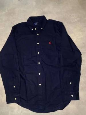 Mörkblå skjorta från Polo Ralph Lauren - Klassisk mörkblå skjorta från Polo Ralph Lauren med button-down krage och vita knappar. Skjortan har lång ärm och den ikoniska röda logotypen broderad på bröstet. Perfekt för en clean och stilren look.   Den är använd 2 gånger. Priset kan diskuteras 