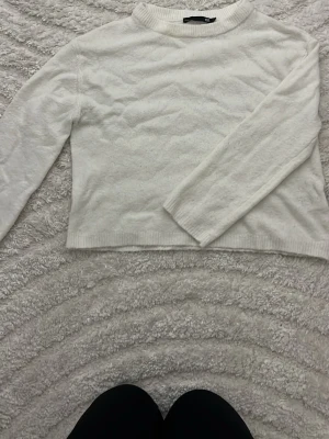 Vit stickad tröja från 157 - Klassisk vit stickad tröja från 157 med rund halsringning och långa ärmar. Tröjan har en enkel och stilren design med ribbade muddar vid hals, ärmslut och nederkant. Perfekt att matcha med jeans eller kjol för en clean look.