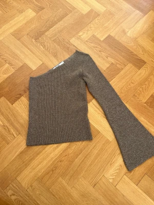 Brun-guldglittrande stickad one shoulder topp - Trendig brun-guldglittrande stickad topp med en axel och lång, utsvängd ärm. Materialet är mjukt och stretchigt, perfekt för dig som vill ha en unik och cool look. Passar till jeans eller kjol för en snygg outfit.