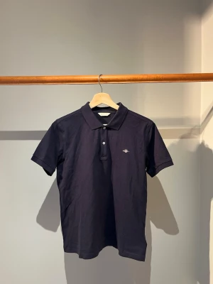 Pikétröja - Klassisk mörkblå pikétröja från Gant med liten broderad logga på bröstet. Tröjan har krage, två knappar framtill och korta ärmar. Perfekt till både jeans och shorts. 