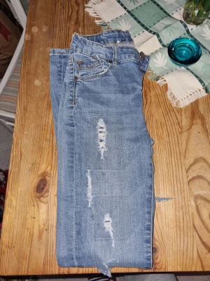 Blå slitna jeans med hål - Säljer ett par blå jeans med trendiga slitningar och hål på benen. Jeansen har klassisk femficksmodell och raka ben. Perfekta för en avslappnad och cool stil. Materialet är denim i bomull. Passar dig som gillar streetwear och en edgy look.