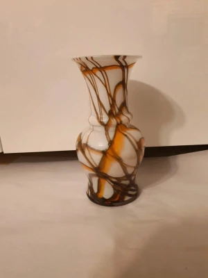 Vit vas med bruna och orange mönster - Unik vas i vitt glas med abstrakta bruna och orange slingor. Vasen har en klassisk form med smal hals och rundad kropp, perfekt som dekoration eller för blommor. Materialet är glas och färgerna ger ett konstnärligt intryck.