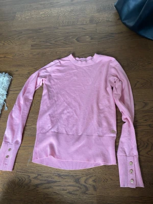 Ljusrosa stickad tröja med knappar - Söt ljusrosa stickad tröja med ribbade muddar och dekorativa guldfärgade knappar vid ärmsluten. Tröjan har rund halsringning och en liten metallbricka vid nederkanten. Perfekt för dig som gillar en feminin och stilren look.