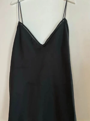 Svart topp med tunna band - En enkel och stilren svart sliptopp med tunna axelband och v-ringning. Toppenhar en rak passform och är tillverkad i ett mjukt, slätt material som ger en snygg siluett. Perfekt att styla lager på lager eller bära som den är.