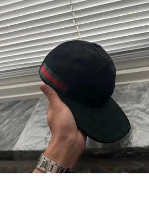 Svart Gucci keps med GG-mönster - Säljer en svart keps från Gucci med klassiskt GG-mönster över hela kepsen. På sidan finns en grön och röd rand som ger den en ikonisk look. Justerbar rem baktill och böjd skärm. Perfekt för dig som vill ha en stilren och lyxig accessoar.