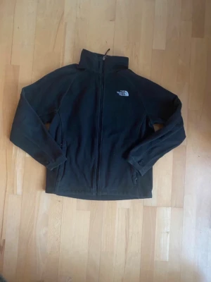 Svart fleecejacka från The North Face - Mysig svart fleecejacka från The North Face med hög krage och dragkedja framtill. Jackan har broderad logga på bröstet och långa ärmar. Perfekt för kyliga dagar och enkel att matcha med olika outfits. Är i väldigt bra skick. Kom gärna med bud