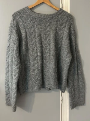 Grå kabelstickad tröja - Mysig grå stickad tröja med klassiskt kabelstickat mönster framtill och på ärmarna. Tröjan har rund halsringning och långa ärmar. Perfekt för kyliga dagar och enkel att matcha med olika outfits.