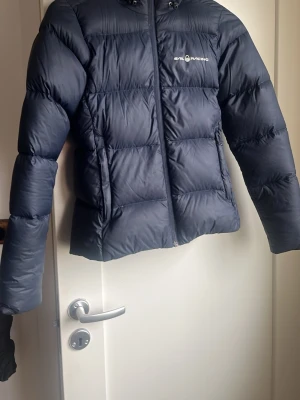 Mörkblå dunjacka Sail Racing - Snygg mörkblå dunjacka från Sail Racing med huva och dragkedja framtill. Jackan har en puffig design med breda kanaler och logga på bröstet samt på huvan. Perfekt för kalla vinterdagar och håller dig varm med sin dunfyllning.