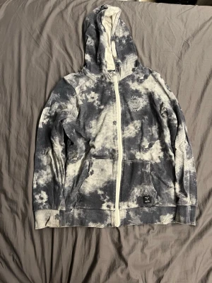 Grå tie-dye hoodie med dragkedja - Säljer en grå och vit tie-dye hoodie från Creative Gymnastics. Hoodien har dragkedja framtill, stor huva och två fickor. Mjuk bomullsmix och coolt tryck på bröstet. Perfekt för dig som gillar streetwear vibes.