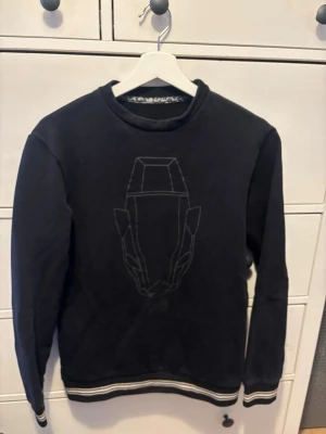 Svart Cavalli sweatshirt - Svart Cavalli sweatshirt med rund hals och broderad märke. Tröjan har ribbade muddar vid ärmslut och nederkant med vita ränder. Perfekt för en avslappnad och stilren look.