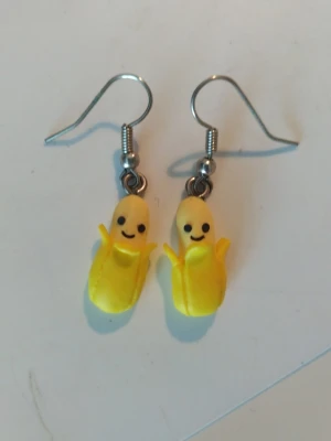 Gula örhängen med glada bananer - Unika, handgjorda örhängen med små glada bananer i starkt gul färg och svarta detaljer för ansikte. Hängena är gjorda i plast och metall, och har krokar i silverfärg. Perfekta för dig som vill ha något lekfullt och färgglatt i öronen.