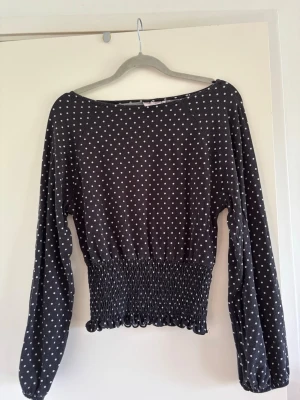 Svart prickig topp - Säljer en svart långärmad topp med vita prickar och smock i midjan. Toppen har en avslappnad passform upptill och sitter tightare vid midjan för en snygg siluett. Perfekt att styla med jeans eller kjol för en trendig look.