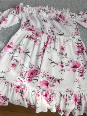 Vit blommig offshoulder klänning - Söt vit offshoulder klänning med rosa och rosa-blommigt mönster. Klänningen har volangdetaljer upptill och nedtill samt långa ärmar. Perfekt för sommarens alla tillfällen och ger en romantisk vibe.