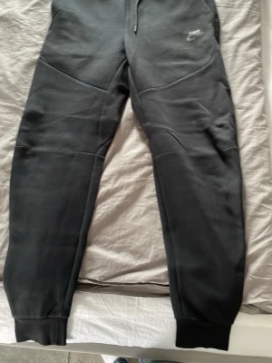 Svarta Nike tech mjukisbyxor - Säljer ett par svarta äkta nike tech mjukisbyxor med broderad logga på vänster lår. Byxorna har dragsko i midjan, muddar vid bensluten och praktiska sidofickor. Tillverkade i mjukt och bekvämt material, perfekta för chill eller träning.   Behöver sälja dessa ASAP🙏