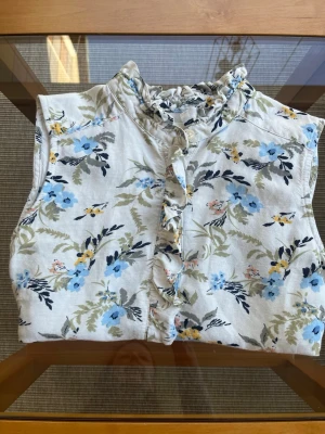 Sö blus med volang från Part Two - Söt ärmlös blus med blommigt mönster i blått, gult och grönt på vit botten. Volangkant längs knäppningen framtill och liten volang vid halsen. 55% linne och 45% viskos. Storlek 34.