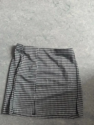 Svartvit rutig kortkjol med slits - Oanvänd snygg svartvit kortkjol med klassiskt rutigt mönster och två små slitsar framtill. Kjolen har en rak passform och är perfekt för dig som gillar en trendig och stilren look.