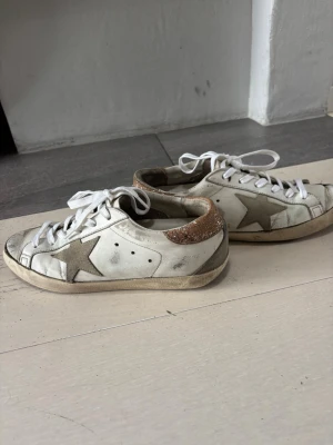 Golden Goose vita sneakers med stjärna - Säljer ett par vita Golden Goose sneakers med grå stjärna på sidan och glitterdetalj i brons på hälen,  med slitna detaljer för en cool vintage-look. Köptes för något år sedan och använda, men fortfarande i relativt bra skick. Jätte bekväma och snygga!! Pris går att diskutera!!