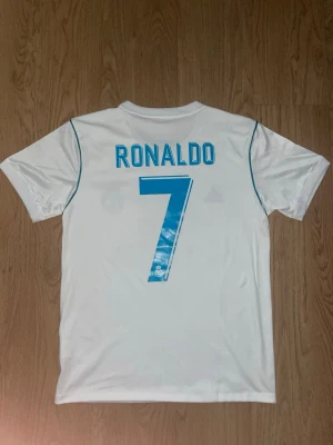 Real Madrid Ronaldo matchtröja - Säljer en vit Real Madrid matchtröja från Adidas med Ronaldo och nummer 7 på ryggen. Tröjan har blå detaljer, klubbmärke och Fly Emirates-tryck på bröstet. Perfekt för dig som älskar fotboll och vill bära din favoritspelare på ryggen.