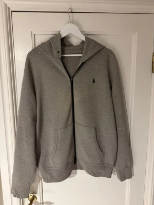 Grå hoodie från Polo Ralph Lauren - Snygg grå hoodie från Polo Ralph Lauren med dragkedja framtill och klassisk logga på bröstet. Hoodien har två stora fickor och ribbade muddar vid ärmar och nederkant. Perfekt för en chill och stilren look.