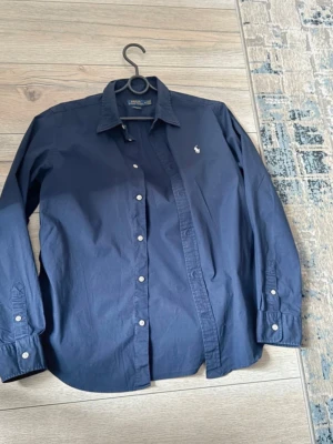 Mörkblå skjorta från Ralph Lauren - Klassisk mörkblå skjorta från Ralph Lauren med vit broderad logga på bröstet. Skjortan har lång ärm, knappar framtill och vid manschetterna samt en tidlös krage. Perfekt till både jeans och chinos.