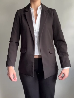 Svart klassisk kavaj med fickor - Stilren svart kavaj med figurnära passform och två fickor med lock framtill. Kavajen har klassiska slag och är helfärgad utan synliga detaljer. Perfekt för en clean och snygg look. Materialet ser ut att vara en mjuk och lätt tygblandning.