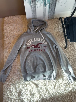 Grå Hollister hoodie med tryck - Säljer en grå hoodie från Hollister med stor magficka och vit dragsko i huvan. På bröstet står det 'Hollister California 1922' i vitt och rött med den klassiska fågel-loggan. Mjuk insida och ribbade muddar. Perfekt för chill dagar.           ‼️Obs ‼️Denna hoodie har krympt efter år av användning , sitter ungefär som S.