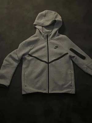 Nike Tech Fleece Hoodie Grå Barnstorlek XL - Nike Tech Fleece-hoodie i grå färg med svarta detaljer. Hel dragkedja framtill, huva och praktiska fickor med dragkedja. En bekväm och stilren hoodie som passar perfekt till vardags, skolan eller träning.