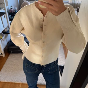 Beige ribbad kofta med knappar - Superfin beige kofta med ribbad midja och silvriga knappar hela vägen framtill. Modellen är figurnära med lång ärm och rund halsringning. 