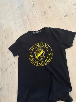 Svart AIK t-shirt med gul logga - Säljer en svart t-shirt från AIK med stor gul klubbmärke och texten 'Allmänna Idrottsklubben' på bröstet. T-shirten är officiell merchandise och har en klassisk passform med rund hals och korta ärmar. Perfekt för dig som vill visa ditt AIK-stöd.