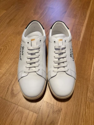 Vita Saint Laurent sneakers - Snygga vita sneakers från Saint Laurent med handskriven logga på sidan. Skorna har klassisk låg profil, vita snören och diskreta svarta detaljer vid hälen. Tillverkade i slätt läder för en clean och stilren look. Perfekta för dig som gillar exklusiva och minimalistiska sneakers.