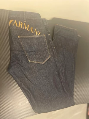 Mörkblå Emporio Armani jeans - Snygga mörkblå jeans från Emporio Armani med tydlig gul logga broderad över bakfickorna. Klassisk femficksmodell med raka ben och kontrastsömmar. Perfekta för dig som vill ha en stilren men ändå statement look. Använt 2 eller 3 gånger bara. Köpt för 1200kr på BESTSECRET 