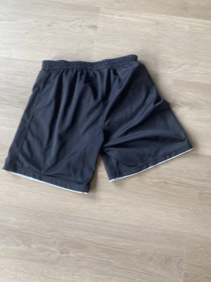 Svarta sportshorts Team Sportia - Svarta sportshorts från Team Sportia med elastisk midja och vit kant längst ner. Tillverkade i ett lätt och svalt syntetmaterial, perfekta för träning eller sport. Klassisk passform och diskret logga på ena benet. 158/164