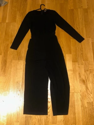 Svart långärmad jumpsuit Bubbleroom - Snygg svart jumpsuit från Bubbleroom med långa ärmar och rund halsringning. Modellen har vida ben och dragkedja bak för enkel påklädning. Perfekt för dig som gillar stilrena plagg med en clean look.