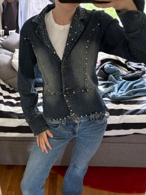 Vintage jeans jacka - Snygg mörkblå jeansjacka med markerad midja och dekorativa silverfärgade nitar längs sömmarna och kragen. Jackan har en unik kavajliknande krage med prickigt innerfoder och stängs med två knappar framtill. Perfekt för dig som vill sticka ut.