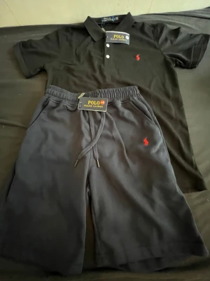 Svart set från Polo Ralph Lauren - Snyggt svart set från Polo Ralph Lauren med pikétröja och shorts. Pikén har klassisk krage, tre vita knappar och röd logga på bröstet. Shortsen har dragsko i midjan och samma röda loggar. Perfekt för en avslappnad och stilren look.