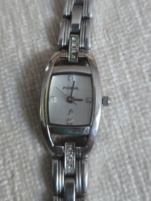 Silverfärgad klocka från Fossil med batteriet  - Stilren klocka från Fossil i silverfärg med rektangulärt ur och länkarmband. Urtavlan har diskreta stenar vid varje kvart och Fossil-logga. Armbandet har dekorativa stenar som ger extra bling. Perfekt accessoar för dig som gillar klassisk men ändå trendig stil.