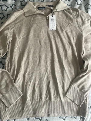 Helt Ny halvzip-tröja Jack & Jones - Snygg beige tröja från Jack & Jones med halv dragkedja och krage. Tillverkad i mjukt material med ribbade muddar vid ärmslut och nederkant. Perfekt för en clean och stilren look. Bara prövad helt ny! 