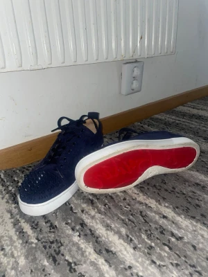 Mörkblå Christian Louboutin sneakers - Snygga mörkblå sneakers från Christian Louboutin i mocka med ikoniska röda sulor och svarta nitar över tån. Skorna har klassisk låg profil, svarta skosnören och en unik edgy look som sticker ut. Perfekta för dig som vill ha något extra till din outfit.