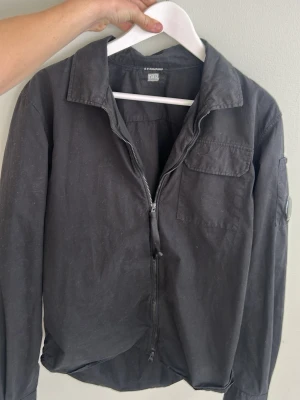 Svart overshirt från C.P. Company - Snygg svart overshirt från C.P. Company med dragkedja framtill, bröstficka med lock och klassisk krage. Tillverkad i ett slitstarkt bomullsmaterial och har en ikonisk linsdetalj på ärmen. Perfekt lager-på-lager-plagg för en cool streetwear-look.