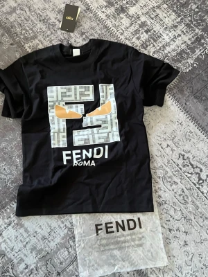 Svart Fendi t-shirt med grafiskt tryck - Snygg svart t-shirt från Fendi med stort grafiskt tryck framtill i vitt, grått och beige samt texten 'Fendi Roma'. Klassisk rund hals och korta ärmar. Perfekt för dig som gillar exklusiva streetwear-plagg.