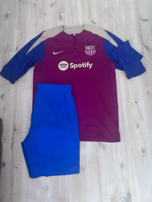 Barcelona träningsset Nike x Spotify - Säljer ett Barcelona träningsset med långärmad tröja i vinrött, beige och blått samt blå shorts. Tröjan har dragkedja vid halsen, klubbmärke och Nike-logga samt Spotify som sponsortryck. Perfekt för fotbollsträning eller chill.