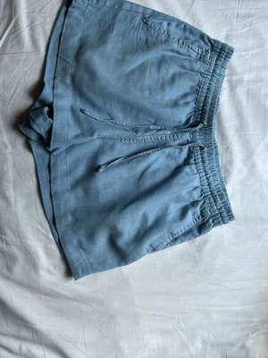 Ljusblå linne shorts med snörning - Snygga ljusblå linne shorts med elastisk midja och snörning framtill. Shortsen har två fickor fram och två dekorativa fickor bak. Perfekta för varma dagar och har en avslappnad passform.