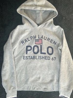 Grå hoodie från Polo Ralph Lauren - Snygg grå hoodie från Polo Ralph Lauren med stor logga och amerikansk flagga på bröstet. Klassisk huva och ribbade muddar vid ärmar och nederkant. Tillverkad i mjuk bomullsblandning för skön känsla. Perfekt för en avslappnad och sportig stil.