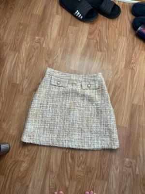 Beige kortkjol från Holly & White - Säljer en beige kortkjol från Holly & White med snyggt strukturerat tyg och två dekorativa fickor framtill med knappar. Kjolen har en klassisk A-linjeform och är fodrad med ljusrosa tyg för extra komfort.