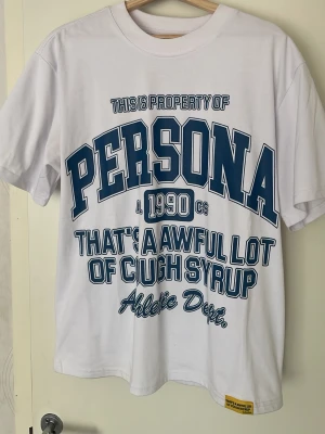 Vit Persona t-shirt med tryck - Säljer en vit t-shirt från Persona med stora blå och röda tryck både fram och bak. Texten inkluderar 'That's a awful lot of cough syrup' och 'Houston Los Angeles'. Klassisk passform och rund hals. Perfekt för dig som gillar streetwear och grafiska prints.