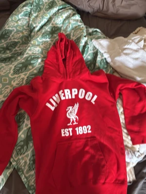 Röd Liverpool hoodie med tryck barn 12/13 storlek  - Snygg röd hoodie med stor vit Liverpool-logga och texten 'EST 1892' på bröstet. Klassisk känguruficka framtill och huva. Perfekt för dig som vill visa ditt stöd för laget och samtidigt hålla stilen. Mjuk och skön bomullsblandning.