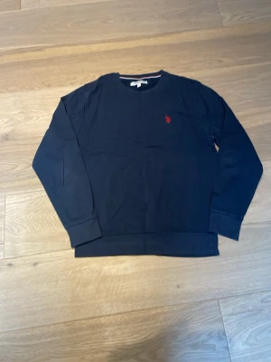 Mörkblå crewneck från Ralph Lauren - Mörkblå långärmad crewneck från Ralph Lauren med röd broderad logga på bröstet. Klassisk passform med ribbade muddar och rund halsringning. Perfekt för en stilren och avslappnad look.