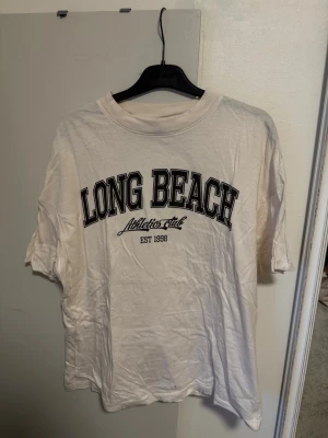 Vit t-shirt Long Beach Divided - Vit t-shirt från Divided med svart text 'LONG BEACH Athletics club EST 1998' på bröstet. Klassisk rund halsringning och korta ärmar. Perfekt för en avslappnad och sportig stil.