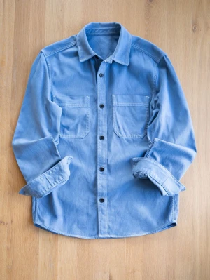Ljusblå overshirt i bomull - Säljer en stilren ljusblå overshirt med två bröstfickor och svarta knappar. Skjortan har klassisk krage, lång ärm och är gjord i mjuk bomull. Perfekt att bära öppen eller stängd för en avslappnad look.